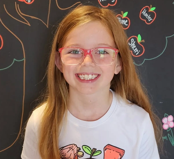 Young girl with red hair and glasses smiling at camera against decorated dark backdrop.