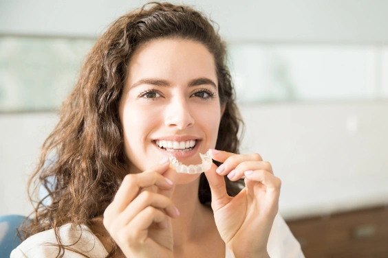 Woman smiling while holding clear aligners in a bright dental office setting.