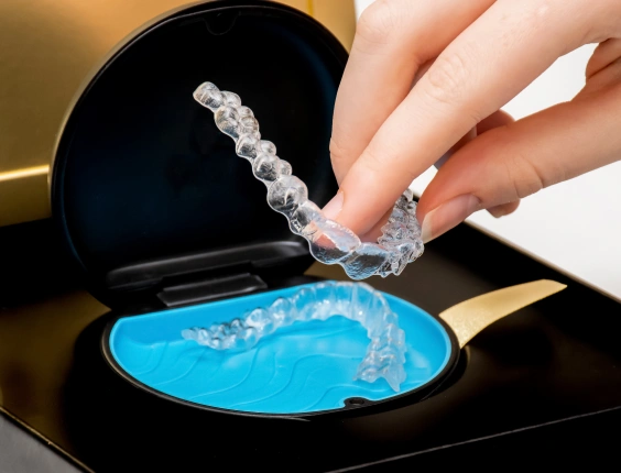 Hand pouring clear aligner into blue orthodontic cleaning solution on black tray.