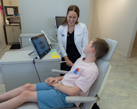 Female orthodontist shows dental scan to young male patient in modern clinic treatment room.
