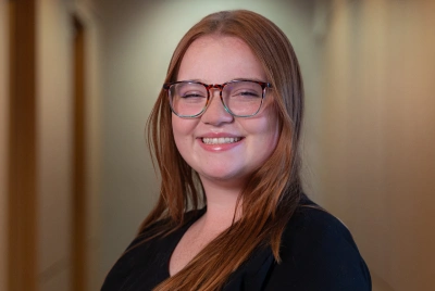 Young woman with red hair and glasses smiling at camera in indoor hallway setting.