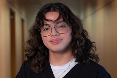 Young person wearing glasses and dark cardigan smiles in indoor hallway setting.