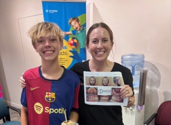 Two women smiling at camera, one wearing Barcelona jersey, holding orthodontics patient photos in clinical setting.