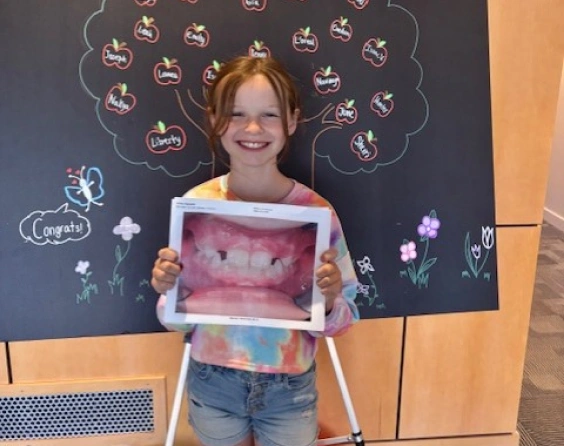 Young girl holding orthodontic treatment photo in front of chalkboard with dental diagrams.