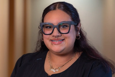 Young woman wearing glasses and black shirt smiles at camera in indoor setting.