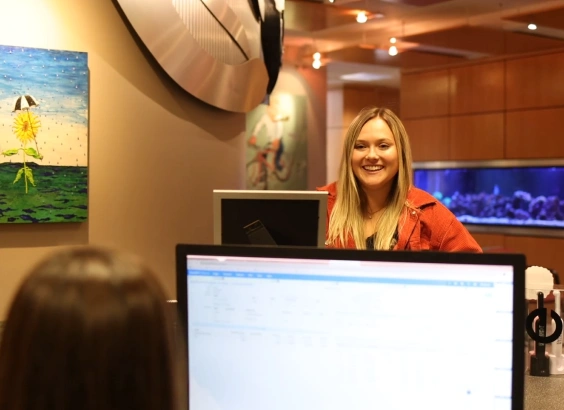 Female receptionist smiling at desk in modern orthodontic office with computer and artwork.