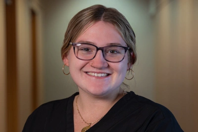 Young woman wearing glasses and black shirt smiling at camera in indoor setting.