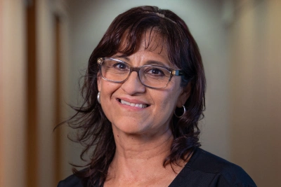 Woman with dark hair and glasses smiling at camera in indoor setting.